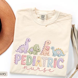Peut inclure: Un t-shirt ivoire avec un motif de quatre dinosaures aux couleurs pastel et le texte PEDIATRIC nurse dans une police style patchwork. Les dinosaures sont violets, verts, roses et bleus, avec des cœurs. Le t-shirt porte une étiquette Comfort Colors.
