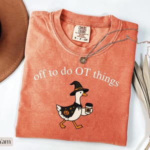 May include: A burnt orange crewneck t-shirt with a graphic of a white goose wearing a black witch hat and cape with a pumpkin print. The text reads off to do OT things in white. The shirt features a Comfort Colors label at the inner neckline.
