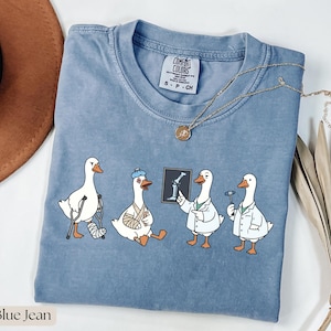 May include: Blue jean coloured t-shirt with a cartoon of four white geese. One goose has a leg cast, another has an arm cast and ice pack, and two are dressed as doctors. A gold necklace is draped on the shirt.