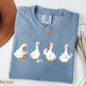 Könnte beinhalten: Blaues Comfort Colors T-Shirt mit einem Cartoon von vier weißen Gänsen mit orangefarbenen Schnäbeln und Füßen. Jede Gans ist in einer anderen Pose, eine hält eine Nagelfeile. Eine goldene Halskette ist über dem Shirt drapiert.