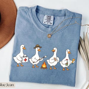 Peut inclure: Un t-shirt bleu jean avec une illustration de quatre canards blancs. Un canard porte une trousse de secours, un autre porte un chapeau et grille une guimauve, un troisième tient un pansement, et le dernier tient un spray et un tube de crème.