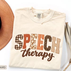 May include: An ivory t-shirt featuring the text SPEECH in block letters with varied patterns including brown plaid, orange polka dots, daisies, orange stripes, leopard print, and tan plaid, with the word therapy in brown cursive script below.