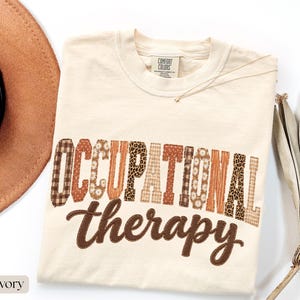 May include: An ivory short-sleeve t-shirt featuring the text "OCCUPATIONAL therapy" in a multi-patterned design. The word "OCCUPATIONAL" is in block letters with various brown, tan, and white prints, while "therapy" is in brown script.