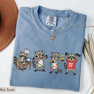 Könnte beinhalten: Blaues Jeans-T-Shirt mit einer Grafik von vier Waschbären in verschiedenen Therapie-bezogenen Szenarien. Ein Waschbär sitzt an einem Schreibtisch, ein anderer benutzt Widerstandsbänder, ein dritter hält Therapieknete und der letzte hebt Gewichte. Das Shirt hat einen Rundhalsausschnitt.