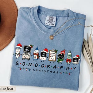 May include: A blue jean colored t-shirt featuring a graphic of cartoon sonography equipment wearing Christmas hats and scarves. The text reads SONOGRAPHY CHRISTMAS with colorful holiday lights. The shirt has a Comfort Colors brand label.