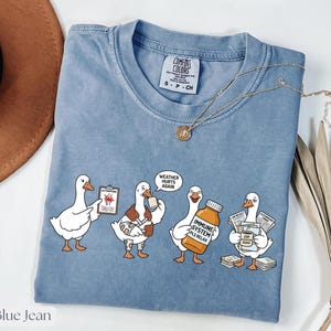 May include: Blue jean Comfort Colors t-shirt featuring a cartoon goose graphic. The graphic depicts four geese in different states of distress, holding items such as a clipboard, a medicine bottle, and newspapers. The text "WEATHER HURTS AGAIN" is in a speech bubble.