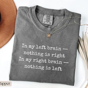 Op de afbeelding: Een grijs Comfort Colors t-shirt met de tekst "In my left brain - nothing is right, In my right brain - nothing is left" in het wit. Een zilveren ketting met een kleine hanger ligt op het shirt. Een bruine hoed is gedeeltelijk zichtbaar.
