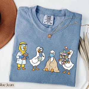 Op de afbeelding: Blauw Comfort Colors T-shirt met een afbeelding van vier cartoon ganzen in medische kleding. Eén gans draagt een beschermingspak, een andere houdt een vergrootglas vast, een derde is in een deken gewikkeld en de laatste draagt medicijnflesjes.