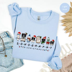 May include: A light blue crewneck sweatshirt featuring a graphic of cartoon sonography equipment wearing Christmas hats and scarves. The text reads SONOGRAPHY CHRISTMAS with colourful holiday lights. The design includes ultrasound machines and gel.