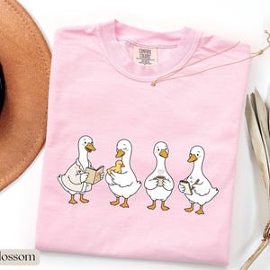 May include: A light pink t-shirt featuring a whimsical illustration of four cartoon ducks. Each duck is depicted with unique accessories: a book, a baby duckling, a coffee cup, and a notepad. The shirt has a crew neck and short sleeves.