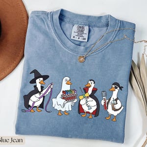 Könnte beinhalten: Ein blaues T-Shirt mit einer Grafik von vier weißen Enten in Kostümen. Eine Ente trägt einen Hexenhut und Umhang, eine ist ein Geist, eine ist ein Vampir mit einer Spritze und eine ist ein Pirat, der ein Reagenzglas und einen Kaffeebecher hält.