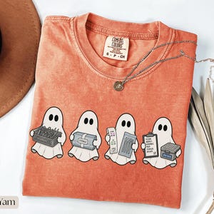 Può includere: Una t-shirt a maniche corte color corallo con una grafica di quattro fantasmi bianchi che tengono in mano forniture mediche. I fantasmi reggono un cestino di strumenti, un pacchetto sterile, una striscia reattiva con strumenti e una cartellina con il testo CLEAN INSPECT ASSEMBLE PACK STERILIZE DELIVER e una scatola etichettata SPD TECH.