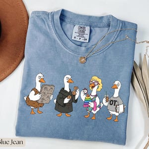 Könnte beinhalten: Blaues Jeans-farbenes Comfort Colors T-Shirt mit einem Cartoon-Gänse-Design. Das Design zeigt vier Gänse in verschiedenen Outfits, die jeweils verschiedene Gegenstände halten. Das Shirt hat einen Rundhalsausschnitt und kurze Ärmel. Der Text "Blue Jean" befindet sich unten links.