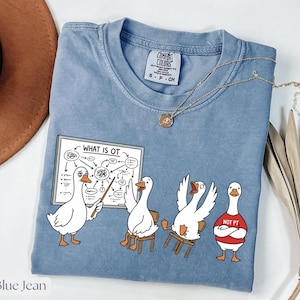 Könnte beinhalten: Blaues Jeans-T-Shirt mit einer Grafik von Cartoon-Gänsen. Eine Gans zeigt auf ein Diagramm mit der Aufschrift "WHAT IS OT". Andere Gänse sitzen auf Stühlen oder tragen ein "NOT PT"-Shirt. Eine goldene Halskette liegt auf dem T-Shirt.