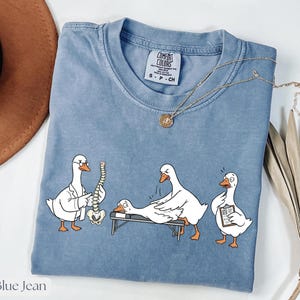 Peut inclure: T-shirt bleu jean Comfort Colors avec un graphisme humoristique de trois oies dans un cabinet de chiropractie. Une oie examine une colonne vertébrale, une autre est sur une table de massage et la troisième tient un presse-papiers. Un collier doré est drapé sur le t-shirt.