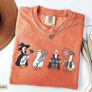 Puede incluir: Una camiseta de color naranja quemado con un gráfico de cuatro gansos disfrazados. Un ganso lleva sombrero de bruja y capa con un martillo de reflejos, otro es un fantasma con un diapasón, otro es un vampiro con una resonancia y otro es pirata.