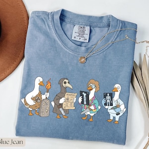 May include: Blue jean colored Comfort Colors t-shirt featuring a humorous graphic of cartoon ducks in various medical and historical roles. The ducks are depicted with a torch, a plague doctor mask, and holding X-rays and medical tools.