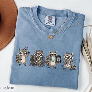 以下が含まれることがあります： クルーネックのブルーデニムカラーのComfort Colors Tシャツ。シャツには、さまざまなストレスのかかったポーズの4匹のアライグマのグラフィックが描かれています。ペンダント付きのゴールドネックレスがシャツにかけられています。シャツには小さなComfort Colorsタグが付いています。