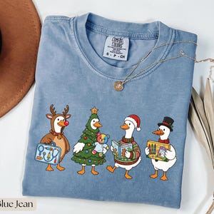 Puede incluir: Una camiseta color azul jean con un gráfico de cuatro gansos blancos vestidos para las fiestas. Un ganso lleva astas de reno y un maletín médico, otro es un árbol de Navidad con un oso de peluche, otro lleva un gorro de Papá Noel y un suéter leyendo un libro, y otro lleva un sombrero de copa con una caja de crayones.