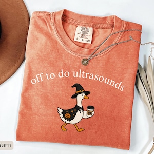 Puede incluir: Una camiseta color ñame con el texto blanco "off to do ultrasounds" sobre un gráfico de un ganso blanco con un sombrero de bruja negro y una capa negra con patrones de calabazas y estrellas naranjas, sosteniendo un café con una calabaza.
