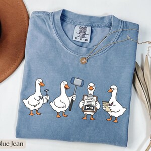May include: A blue jean colored t-shirt featuring a graphic of four white geese. One goose holds a small hammer, another a large mallet, the third holds a clipboard labeled "OFFICIAL ORTHO PLAN: HAMMER, BIGGER HAMMER", and the fourth holds a notepad.