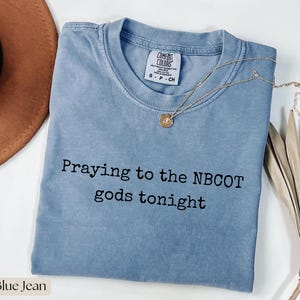 May include: A blue jean colored short-sleeve t-shirt with a crew neckline and a Comfort Colors brand label. The shirt features the black printed text "Praying to the NBCOT gods tonight" centered on the chest. A gold-toned necklace rests on the fabric.