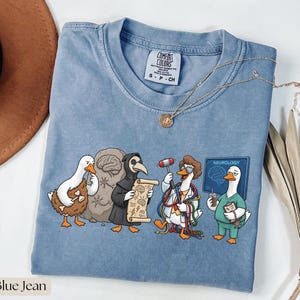 Könnte beinhalten: Blaues Comfort Colors T-Shirt mit einem skurrilen Design von Cartoon-Enten in verschiedenen medizinischen Rollen. Die Enten werden als Höhlenmensch, Pestdoktor, Arzt mit Hammer und Neurologe dargestellt. Das Wort "Neurologie" steht auf einem Schild.