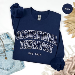 Puede incluir: Una sudadera azul marino de cuello redondo con letras blancas de estilo universitario que dicen OCCUPATIONAL THERAPIST y EST 2027. La prenda de manga larga tiene cuello, puños y dobladillo acanalados con una etiqueta de marca visible.