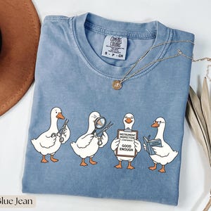 May include: A blue jean colored t-shirt featuring a graphic of four white geese performing tasks. One goose holds scissors, one uses a magnifying glass, one holds a clipboard labeled INSTRUMENT INSPECTION GOOD ENOUGH, and one reads a book.