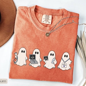 Peut inclure: Un t-shirt à manches courtes orange brûlé avec un motif de quatre fantômes blancs. Le premier fantôme tient une sonde d'échographie, le deuxième une machine, le troisième une image d'échographie et du gel, et le quatrième porte un badge.