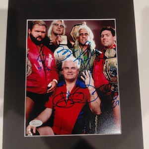 May include: A framed photograph featuring a group of five men, likely wrestlers, posing together. They are wearing colorful outfits and championship belts. The photo is signed in blue ink and includes a JSA authentication sticker.