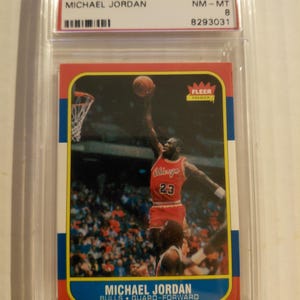 Puede incluir: Una tarjeta de baloncesto de Michael Jordan de 1986 de Fleer en una funda protectora. La tarjeta presenta a Michael Jordan con un uniforme rojo de los Chicago Bulls, en el aire, a punto de encestar. La tarjeta está etiquetada "#57 NM-MT 8".