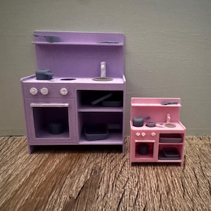 May include: A large lavender miniature kitchen stove unit with a matching smaller pink miniature kitchen stove unit. Both units feature a stovetop, an oven, open shelving, and a backsplash, accompanied by small gray cookware accessories.