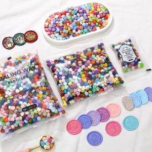 Peut inclure: Un assortiment de perles de cire à cacheter et de sceaux de cire colorés. L'image montre des sachets de perles de cire, un plat ovale blanc rempli de perles et divers sceaux de cire finis de différentes couleurs et de différents motifs. Le texte "SEALING WAX PARTICLES" est visible.