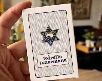 Może przedstawiać: Biała karta z czarnymi i niebieskimi liniami. W środku znajduje się niebieska gwiazda z złotym okiem. Tekst "Estrella Lenormand" jest nadrukowany złotymi literami.