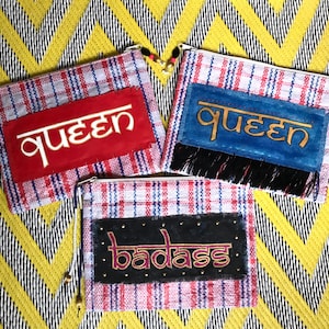 May include: Three colorful zippered pouches with a plaid pattern. The pouches have different embroidered designs: "queen" in gold on red, "queen" in gold on blue, and "badass" in gold on black.