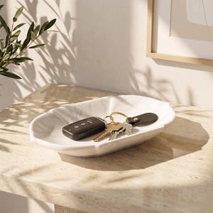 Peut inclure: Un plateau ovale en céramique blanche avec un bord festonné, présentant un motif subtil rappelant le marbre gris. À l'intérieur du plateau repose une clé de voiture noire attachée à un anneau en métal doré avec des clés argentées et une étiquette en cuir noir.