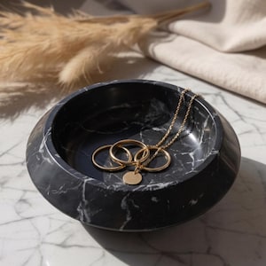 Puede incluir: Un cuenco redondo de mármol negro con vetas blancas que contiene tres anillos de metal dorado y un collar de cadena fina dorada con un pequeño colgante circular que descansa dentro del cuenco.