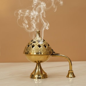 Pode incluir: Um queimador de incenso de latão com uma tampa decorativa perfurada e uma alça. Volutas de fumaça branca sobem do topo, sugerindo que o queimador está em uso. O queimador tem uma base de pedestal e fica em uma superfície clara.