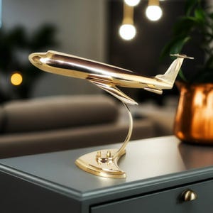 May include: A gold-colored airplane model, displayed on a curved stand. The model has a polished finish, reflecting light. The airplane is a decorative item, likely for display on a desk or shelf. The stand is attached to a dark gray surface.