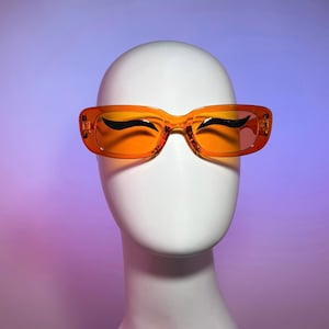 Puede incluir: Gafas de sol rectangulares naranjas con detalles curvos negros, exhibidas sobre una cabeza de maniquí blanca. Las gafas tienen una montura y lentes naranjas transparentes. El fondo es un degradado de morado y rosa.