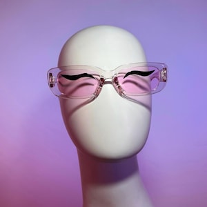 Puede incluir: Gafas de sol rectangulares transparentes con lentes rosas y detalles negros estilo delineador. Las gafas se muestran sobre una cabeza de maniquí blanca sobre un fondo degradado púrpura y rosa. Diseño retro y elegante.