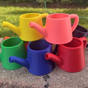 May include: A collection of small, matte-finish watering cans in vibrant colors including yellow, pink, green, blue, red, and purple. Each miniature watering can features a simple cylindrical body, a side handle, and a short, narrow spout.