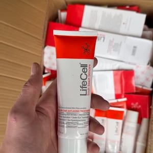 Op de afbeelding: Een witte en oranje tube LifeCell South Beach Skincare ALL-IN-ONE ANTI-AGING TREATMENT wordt in een hand gehouden. De tube heeft een rode bovenkant en een witte basis, met de merknaam en productdetails erop gedrukt. Andere soortgelijke tubes zijn zichtbaar op de achtergrond.