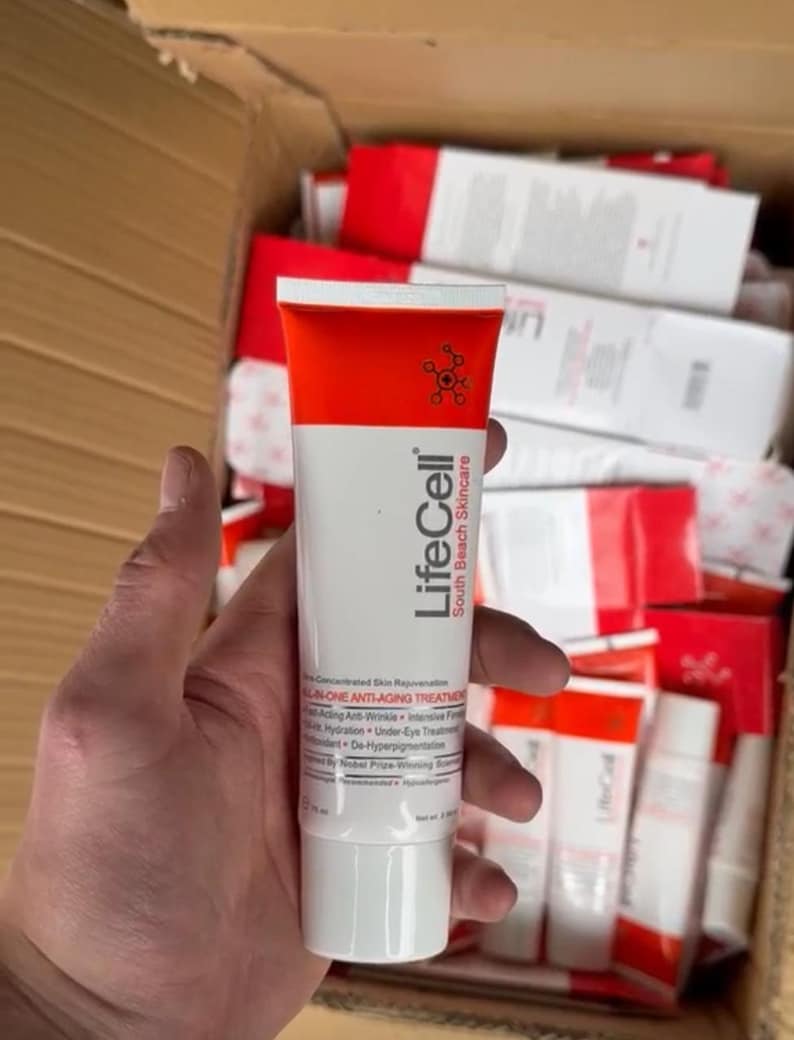 Op de afbeelding: Een witte en oranje tube LifeCell South Beach Skincare ALL-IN-ONE ANTI-AGING TREATMENT wordt in een hand gehouden. De tube heeft een rode bovenkant en een witte basis, met de merknaam en productdetails erop gedrukt. Andere soortgelijke tubes zijn zichtbaar op de achtergrond.
