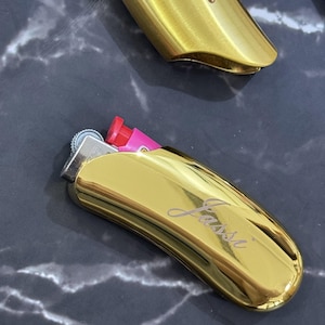 以下が含まれることがあります： 光沢のあるゴールドトーンの金属製ライターケースで、曲線的で人間工学に基づいた形状をしています。ケースの前面には「Jassi」という筆記体のテキストが刻印されています。ピンク色のライターが上部に部分的に挿入されており、銀色の着火ホイールが見えています。