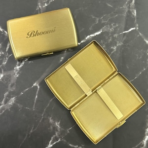 May include: A gold-toned metal cigarette case with a brushed finish. One case is closed and features the word Bhoomi engraved on the front. The other case is open, revealing a textured interior with two metal spring-loaded bars to hold items in place.