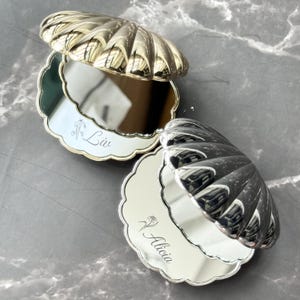 May include: Two clam shell-shaped compact mirrors with metallic finishes. One mirror is gold and features the name Liv with a small flower engraving. The other mirror is silver and features the name Alicia with a small flower engraving. Both mirrors have scalloped edges and reflective surfaces.