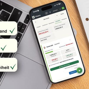 May include: A smartphone displaying a financial app interface, resting on a laptop keyboard. The screen shows account details and financial data. Three labels with German text are visible: "Aktueller Stand", "Restbetrag", and "Schuldenfreiheit". A notepad and pen are in the background.
