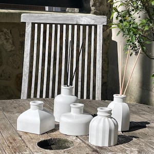 Peut inclure: Cinq flacons diffuseurs de parfum en céramique blanche de formes variées, certains avec des bâtonnets. Les flacons sont posés sur une table en bois vieilli, avec une chaise en bois et de la verdure en arrière-plan. Les flacons sont vides.